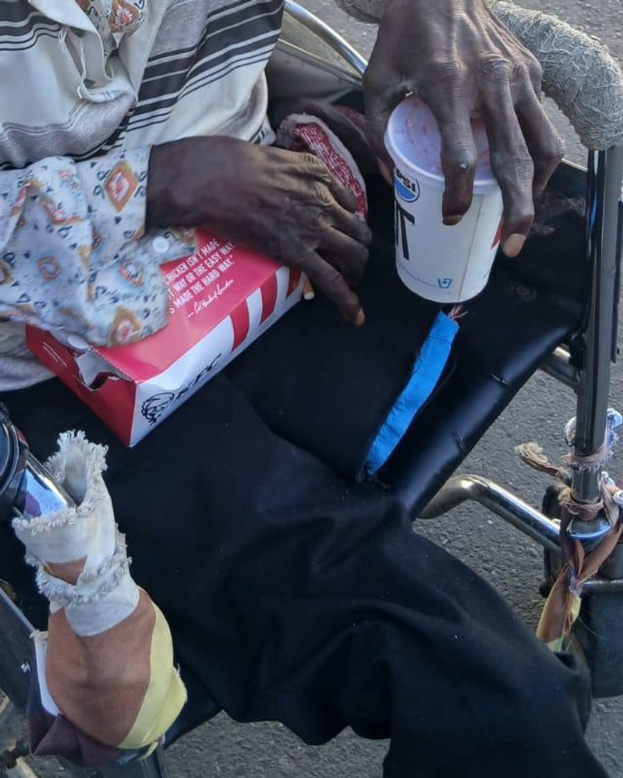 Bandaged hands resting on a drink and a boxed meal, in a wheelchair at the roadside.